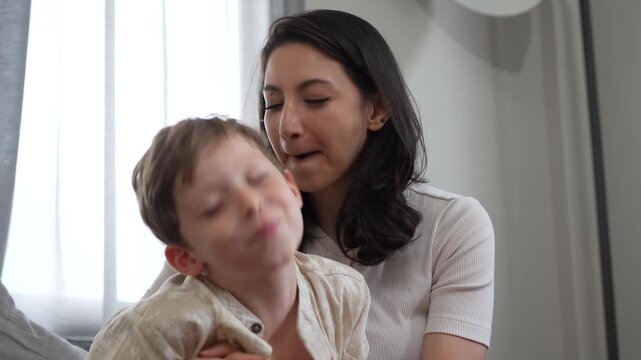 Son kissing mother on cheek and hugging at home, little boy embracing and kissing happy mother indoors, mother and son sharing tender moment and affection on sofa