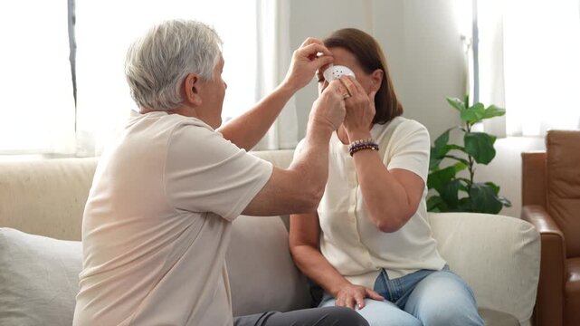 Senior man helping wife applying eye patch at home, elderly husband taking care of wife with eye bandage indoors, senior couple healthcare support and medical eye care at home