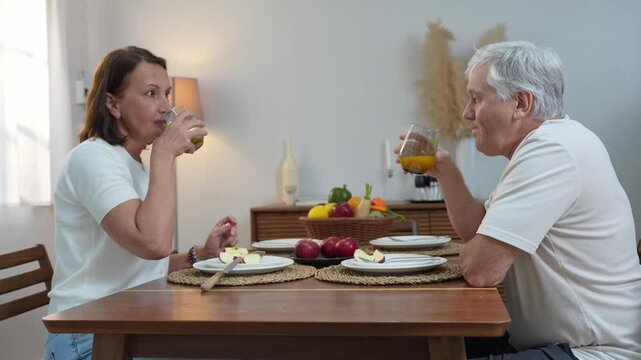 Senior couple toasting with juice during breakfast at home, Happy retired couple enjoying healthy drink together at dining table, Elderly man and woman celebrating with fruit juice at dining room