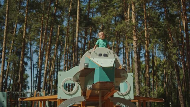 Wide shot of a young boy standing on top of a large spaceship-themed wooden playground structure in a forest. Active child exploring a massive outdoor play complex in a park.