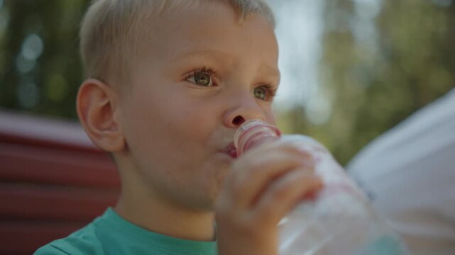 Kid drinking water outside.  Close-up portrait of a young blonde boy drinking fresh water from a plastic bottle outdoors. Child taking a refreshing drink break from playing in a sunny outdoor park.