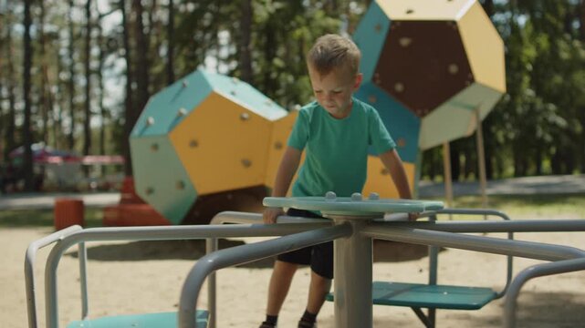 Boy playing on a merry-go-round carousel at a sunny public playground. Happy child spinning on an outside playground carousel. Baby enjoying activities in a park outdoors.