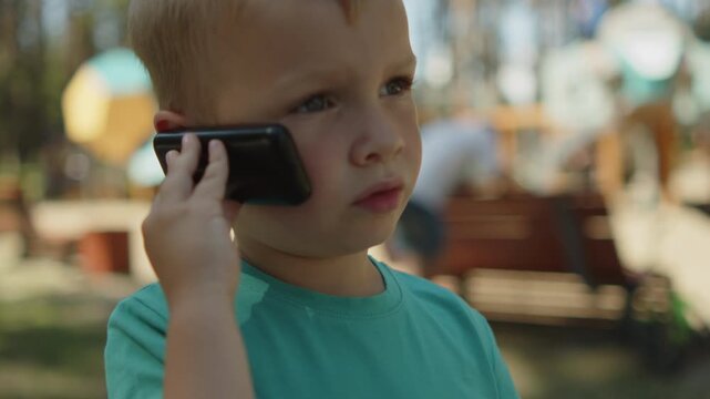 Kid talking on an old telephone outside. Young boy holding a black phone to his ear. Child speaking by a cellular phone device in a park. Small baby using a small phone in a park.