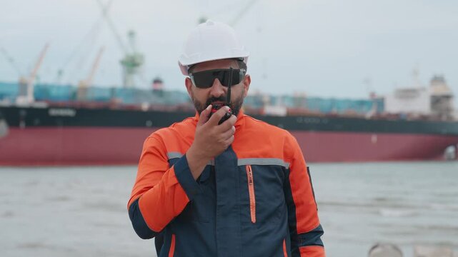 Worker stands near water, talking on a walkie-talkie. He wears a helmet and sunglasses. Large ship looms in the background