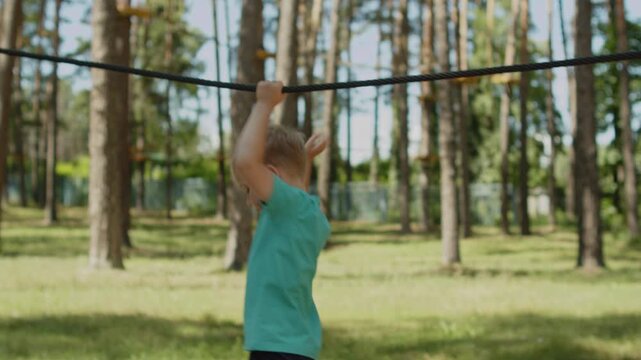 Young blonde boy on an outdoor slackline course in a sunny forest park. Child playing on a playground outside. Small kid doing activities outdoors. Happy kid on an obstacle course.