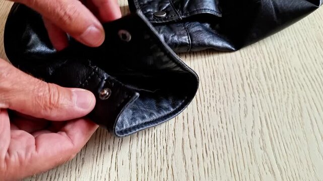 Hands inspect the cuff and inner seam of a black leather jacket on a wooden table indoors. Fashion detail with leather texture, stitching, garment care, and everyday clothing maintenance