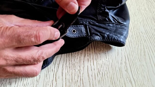 Finger points to the buckle and strap detail on a black leather jacket indoors. Fashion close up highlights leather texture, metal hardware, and garment detail in soft natural light