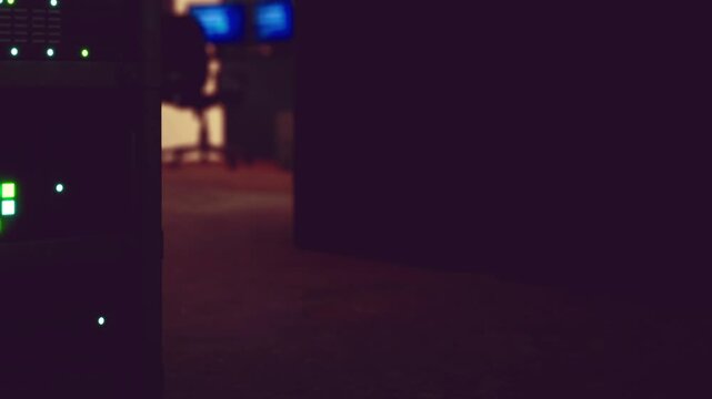 A server rack stands in a dimly lit room. Computer screens glow in the background. The scene shows a workspace likely for tech activity at night.