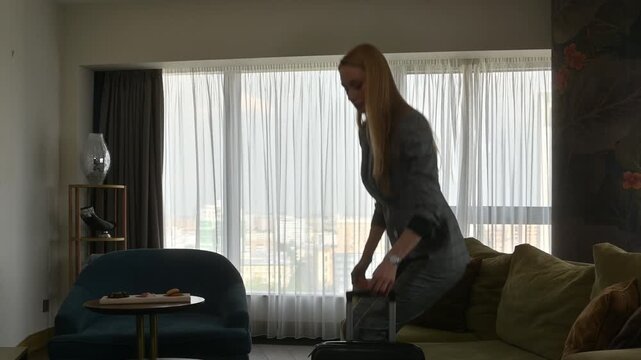Businesswoman in a suit walking into her hotel room with a suitcase, sitting down on the sofa to rest after her arrival, looking content with her luxurious accomodations