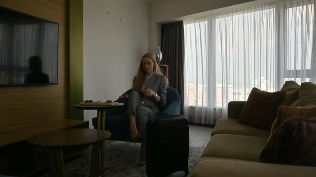 Young businesswoman arriving in her modern hotel room with a suitcase, sitting down in an armchair, and immediately starting to use her smartphone after her business trip