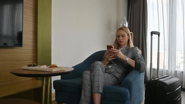 Businesswoman relaxing in an armchair while texting on her mobile phone after checking into her hotel room, with her suitcase standing nearby and sweets on the table