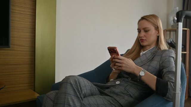 Businesswoman in a plaid suit sitting on an armchair in her hotel room, messaging on her smartphone with a suitcase standing nearby after checking in for a business trip