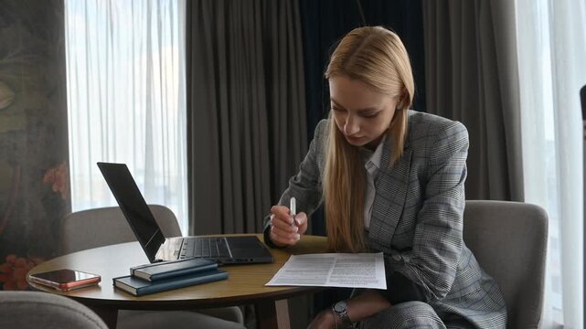Businesswoman with long blonde hair, wearing a formal suit, is signing a contract while sitting at a table with her laptop and notebook in a hotel room during a business trip