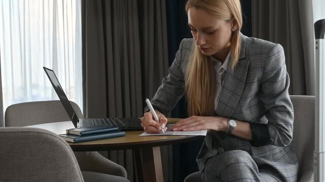Businesswoman in a gray suit sitting at a table in a hotel room, writing on paper with a pen, preparing for a business trip meeting with her laptop and luggage nearby