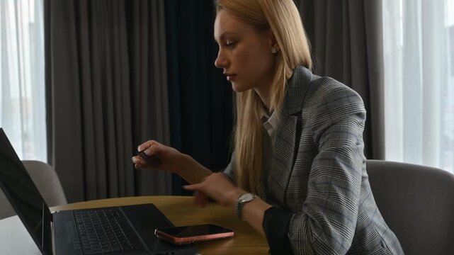 Young woman in a suit sitting at a table in a hotel room, entering her credit card details on a laptop to make an online purchase or book a service while on a business trip