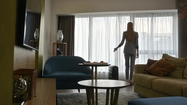 Woman in a stylish suit pulling her suitcase, entering a spacious and elegant hotel suite and approaching the large window to admire the city view, rear view