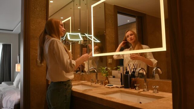 Young woman with long hair brushing it in front of a mirror with integrated light in a modern bathroom, reflecting on her daily routine and personal care concepts