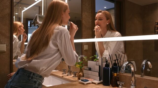 Young woman with long blonde hair applying lipstick while looking at her reflection in a lighted bathroom mirror, getting ready for the day with her daily beauty routine