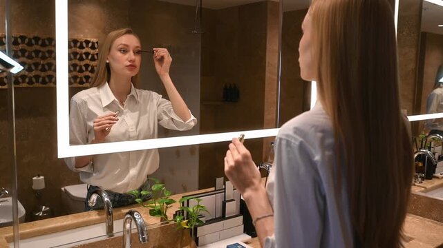 Young woman with long blonde hair applying mascara to her eyelashes, getting ready for the day while looking at her reflection in a hotel bathroom mirror