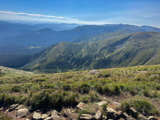 Fototapeta premium Wide alpine hiking trail descends over a vast mountain valley with blue sky and distant ridges, ideal for travel, trekking and outdoor adventure themes.