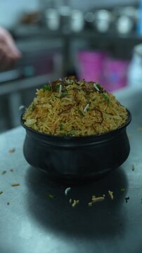 Vertical video close up of chef hands sprinkling finely chopped green coriander over hot dum biryani in black clay pot