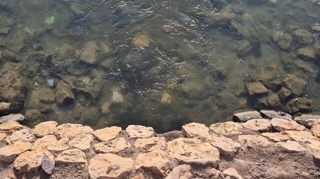 High angle shot of clear flowing water with rocks on riverbed and stone retaining wall embankment