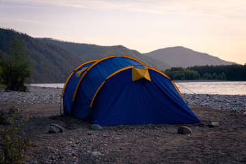 Blue and yellow dome tent set on rocky riverbank with forested hills and calm water at sunrise © Mikhail Mishchenko