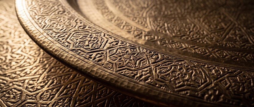 Engraved brass tray with geometric islamic patterns reflects warm golden light across textured metallic surface for cultural display.