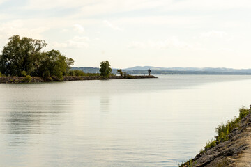 Fototapeta premium Paysage paisible d’un lac aux eaux calmes, bordé d’arbres et de végétation, avec une rive et des reliefs à l’horizon sous un ciel lumineux.