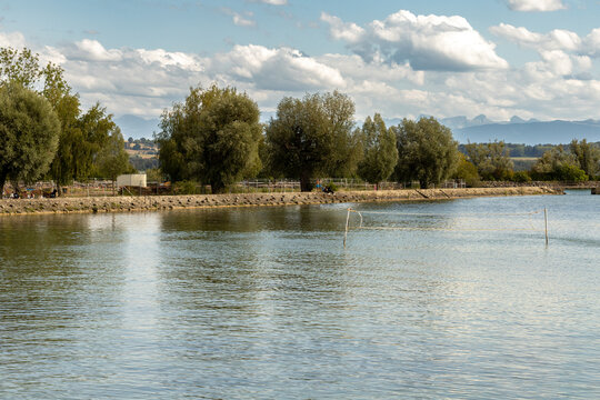 Paysage paisible d&rsquo;un lac aux eaux calmes, bord&eacute; d&rsquo;arbres et de v&eacute;g&eacute;tation, avec des montagnes et des nuages &agrave; l&rsquo;horizon.