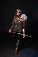 Fototapeta premium Young Viking woman with combat makeup on her face in chainmail with fur and a sword in her hands posing while sitting on a dark background.