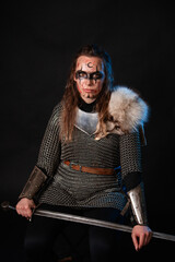 Fototapeta premium Young Viking woman with combat makeup on her face in chainmail with fur and a sword in her hands posing while sitting on a dark background.