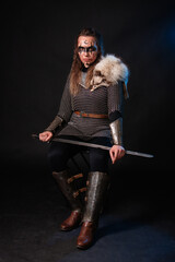 Fototapeta premium Young Viking woman with combat makeup on her face in chainmail with fur and a sword in her hands posing while sitting on a dark background.