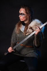 Fototapeta premium Young Viking woman with combat makeup on her face in chainmail with fur and a sword in her hands posing while sitting on a dark background.
