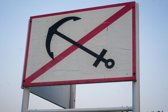 Close-up low angle view of white square maritime sign with red border and crossed out anchor symbol against clear dusk sky background.