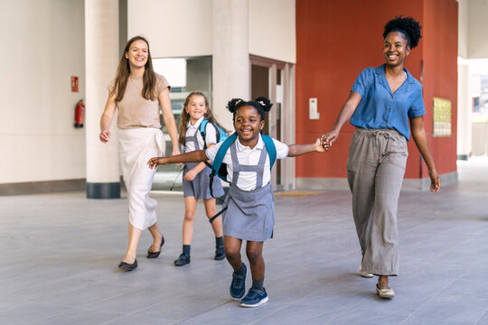 Mothers walking hand in hand with their happy multiethnic girls, wearing school uniforms and backpacks, on their first day back to school