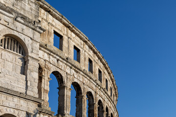 Obraz premium Pula Arena Roman Amphitheater in Croatia. Historic Pula Arena in Croatia with ancient Roman stone arches and curved amphitheater facade illuminated by sunlight beneath a clear blue sky.