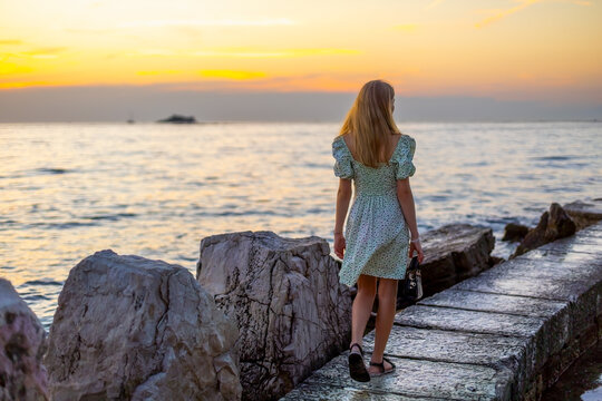 Woman Walking on Rocky Seaside Pier at Sunset. Teenage girl in a floral dress walking along a rocky seaside pier at sunset, carrying a black handbag beside calm ocean water.