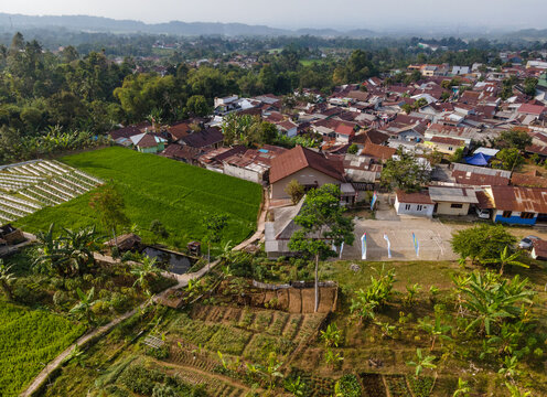 Aerial View of Rice Fields Adjacent to a Rural Settlement with Green Hills in the Distance. Scenic Agricultural Landscape Reveals the Harmony Between Farmland, Villages, and Highland Environment.