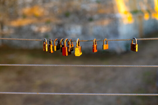 Colorful Love Padlocks Hanging on Steel Cable at Sunset. Colorful weathered padlocks hanging from a steel cable in warm golden sunlight with a softly blurred stone background.