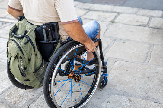 Person Using Manual Wheelchair with Backpack on Stone Plaza. Rear view of a person using a manual wheelchair with a green backpack while moving across a stone plaza.
