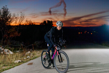 Naklejka premium Female Cyclist Riding Road Bike At Dusk On Empty Country Road With Helmet And Lights