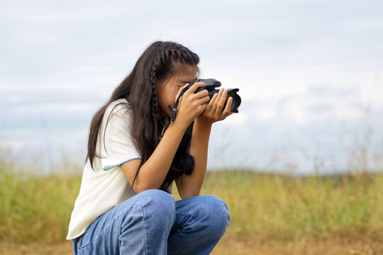 Young girl photographer squatting outdoors taking photos with a DSLR camera against sky background.