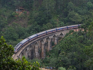 Fototapeta premium Iconic Nine Arch Bridge with Blue Train Crossing in Ella, Sri Lanka. Majestic Colonial Stone Viaduct Architecture Surrounded by Lush Green Tropical Jungle Forest Landscape 