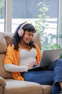 Smiling young woman with curly hair wearing pink headphones, working on laptop while sitting in armchair at home, cozy lifestyle and remote work concept