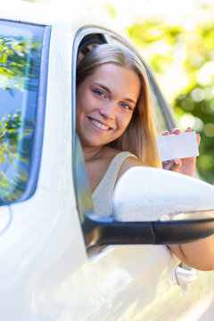 Happy young woman leaning out of car window smiling and showing her new driver's license, celebrating passing driving test outdoors