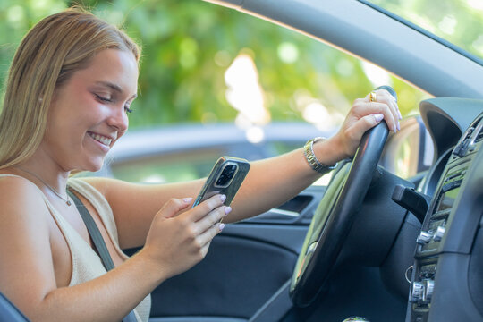 young woman using smartphone while sitting behind the wheel of a car, one hand resting on the steering wheel, texting and browsing on her phone before or while driving