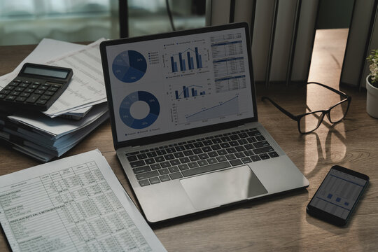 Financial data analysis dashboard on laptop beside calculator, printed reports, smartphone, eyeglasses, showing business performance review, accounting reporting workflow.