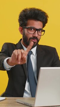 Indian businessman working on laptop shakes finger and saying No be careful scolding and giving advice to avoid danger mistake disapproval sign at office. Confident freelancer man on yellow background