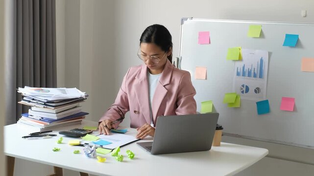 Asian businesswoman holding head on desk with crumpled paper notes, laptop and document stack for corporate financial stress, overworking problem, burnout and fatigue concept